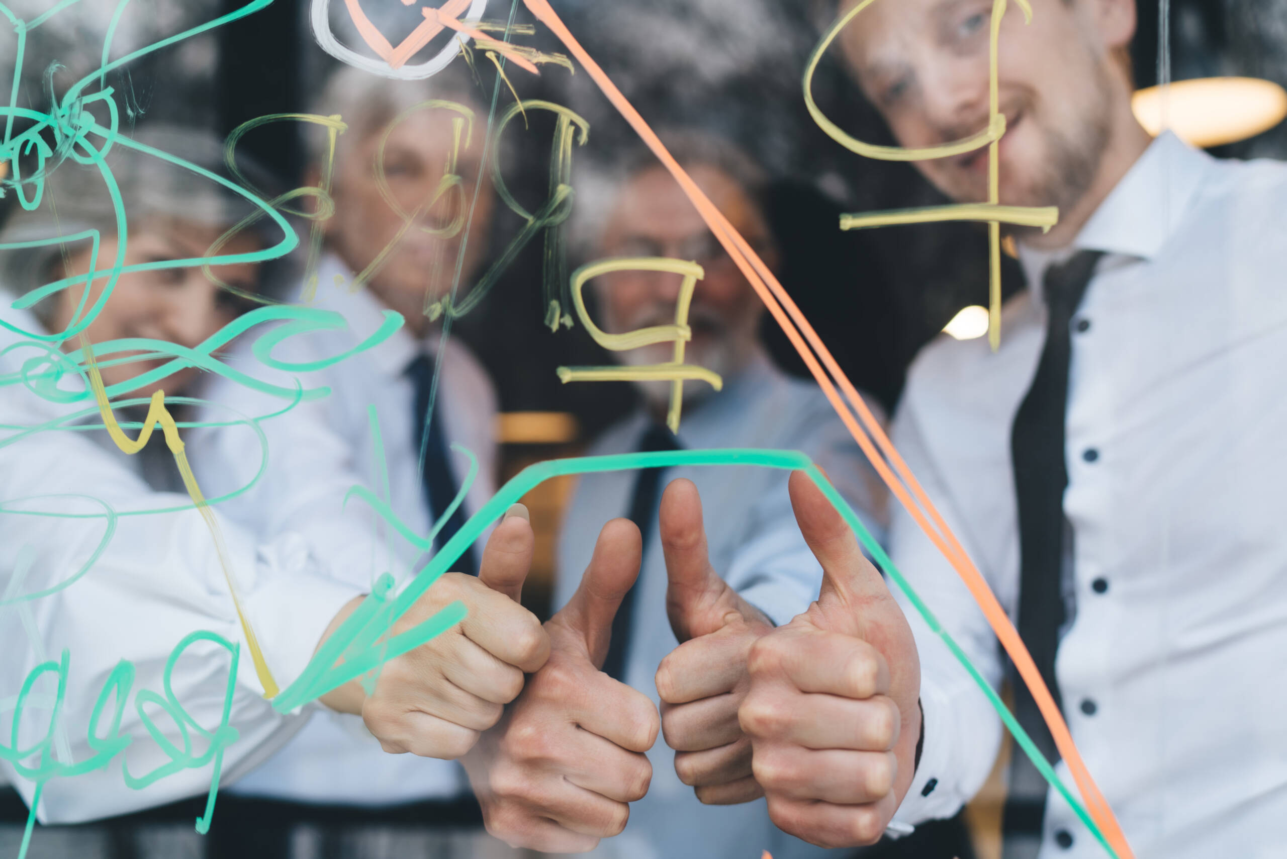 Group of crop cheerful colleagues wearing formal clothes looking at camera while standing near transparent board and showing thumbs up